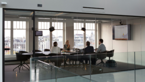 A modern glass-enclosed meeting room with four professionals engaged in a discussion around a dark table. The room features a large screen displaying a presentation, pendant lights, and a cityscape visible through the windows. The setting emphasizes collaboration and technology, aligning with the theme of Dynamics 365 ERP solutions.