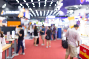 Blurred image of a bustling indoor trade show with attendees walking on a red-carpeted floor, surrounded by brightly lit booths and displays, capturing the dynamic and energetic atmosphere of trade shows.