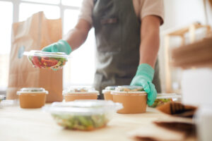 Close-up of a food service worker wearing gloves, carefully packaging fresh salads into eco-friendly brown bowls with clear lids on a light wooden countertop. A brown paper bag for takeout is visible in the background, emphasizing sustainable food delivery packaging.