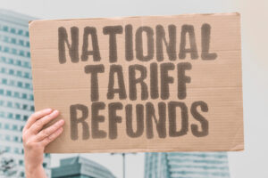 A person holds a cardboard sign reading 'NATIONAL TARIFF REFUNDS' against a blurred urban backdrop, symbolizing advocacy for small business tariff refunds.