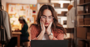 A small business owner in a creative workshop reacts with concern while looking at her laptop, surrounded by clothing designs, shipping boxes, and tools of her trade, symbolizing the challenges of small business crisis communication.