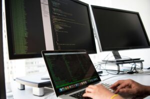 Multi-monitor setup displaying a code editor and a laptop terminal interface operated by expert managed IT providers.