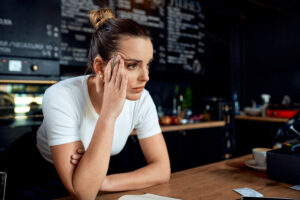 A stressed small business owner stands behind the counter of a cafe, reviewing paperwork near a point-of-sale terminal, illustrating concerns around small business optimism and the latest NFIB outlook.